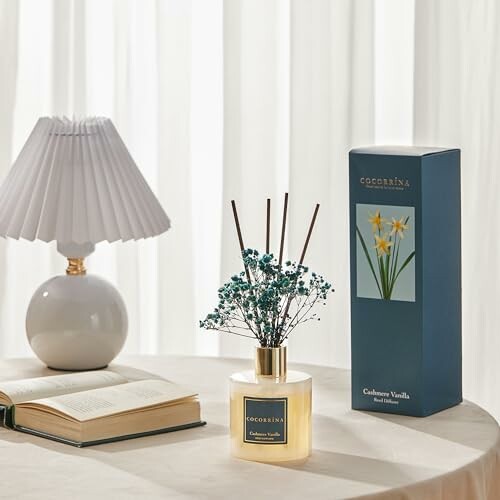 Reed diffuser with floral decoration, lamp, and book on a table.